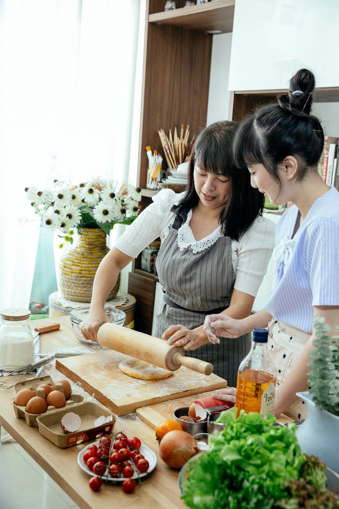 Mother in apron teaching daughter rolling dough for making homemade noodles on kitchen table with vegetables herbs eggs and cooking ingredients