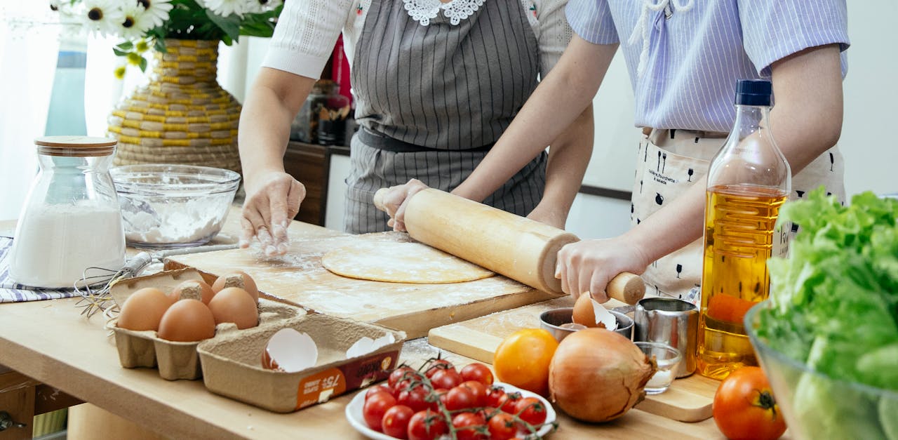 Unrecognizable female cooks rolling dough with pin on wooden board at table with various products while preparing pastry in kitchen