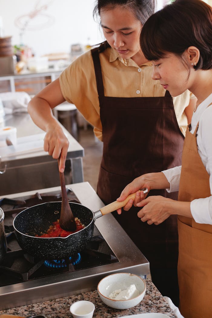 Two women wearing aprons cook together in a modern kitchen, focusing on a pan dish.
